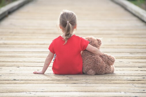 A young girl in a red shirt sitting on a wooden bridge holding a teddy bear, looking away from the camera.